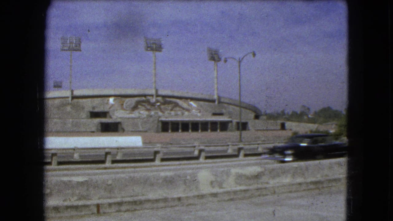 Stadium exterior with floodlights and mural seen across a highway.