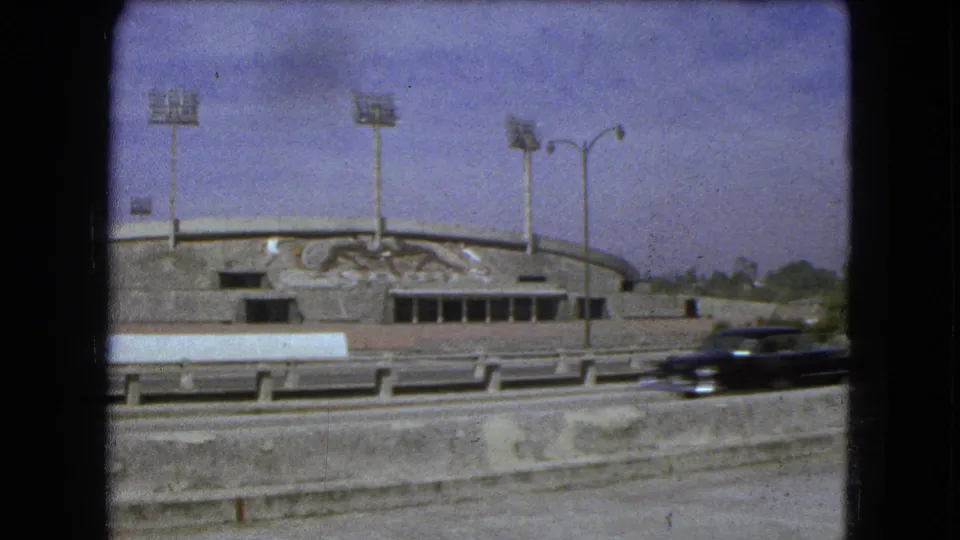 1962 Mexico City 8mm frame showing a concrete stadium facade with floodlights, mural, guardrail, and a passing dark car.