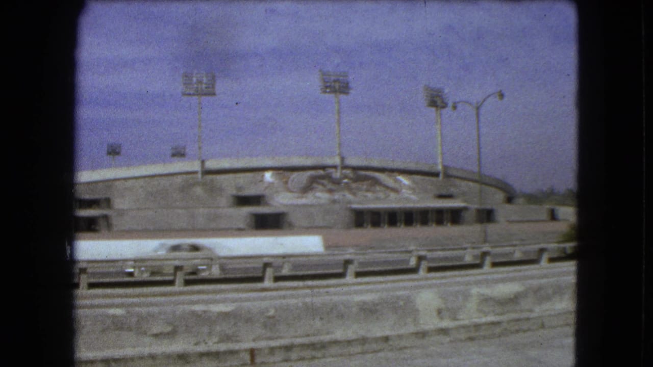 Concrete stadium facade with floodlights, mural, and highway traffic.