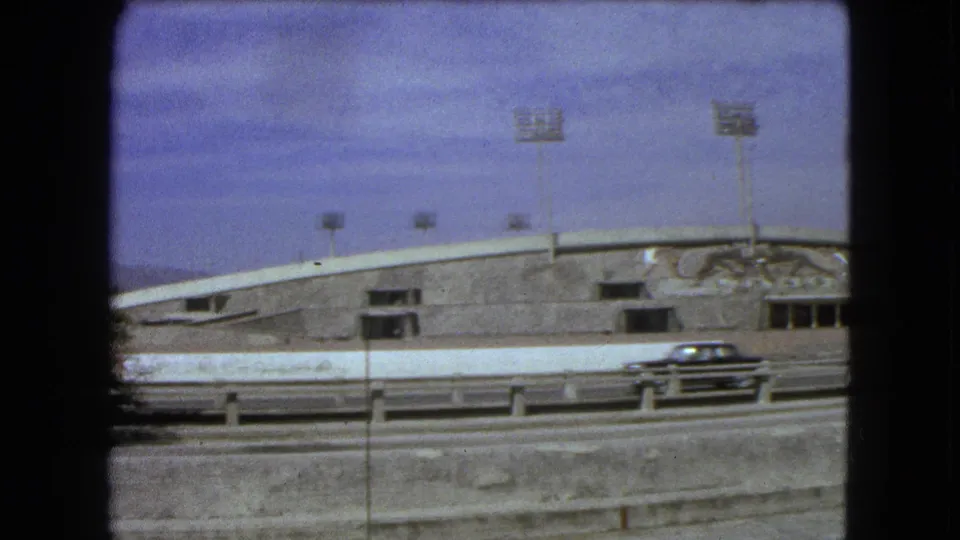 1962 Mexico City 8mm frame showing a dark car on the road below a concrete stadium exterior with roofline lights and mural detail.