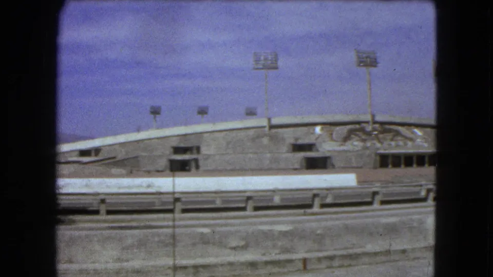 1962 Mexico City 8mm frame with a concrete stadium-like facade, floodlight towers, mural detail, streetlight, and highway barriers.