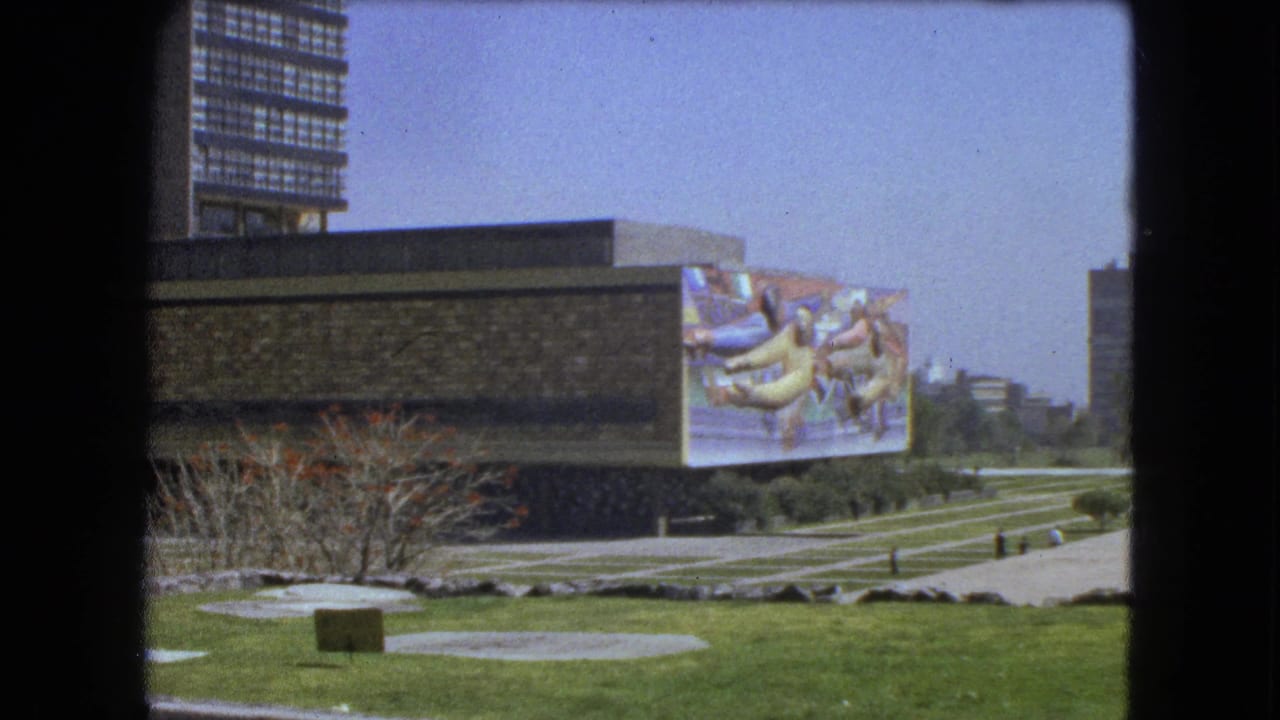 Modern stone venue beside a lawn, walkway, mural, and tall city buildings.