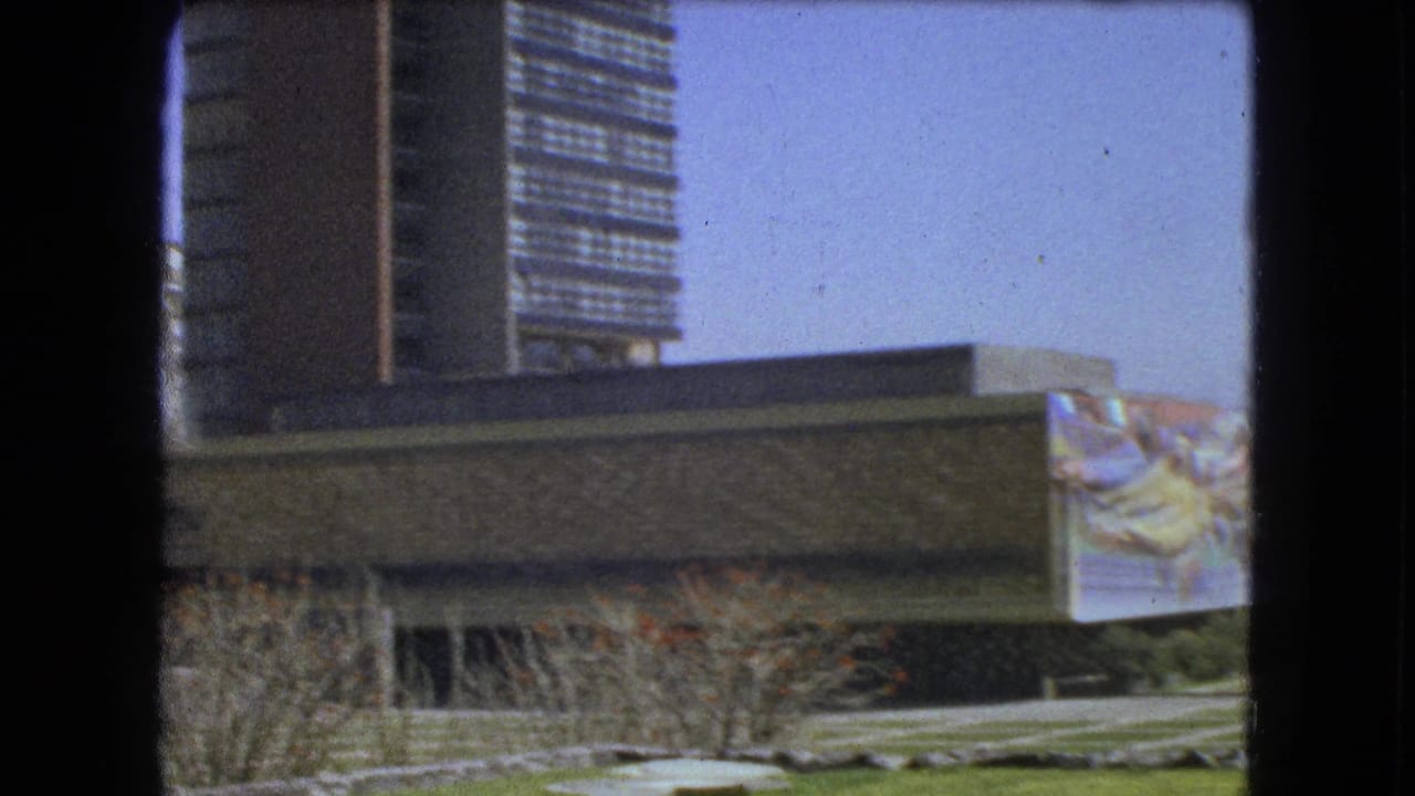 Stone building with mural panel, open lawn, paths, shrubs, and high-rise background.