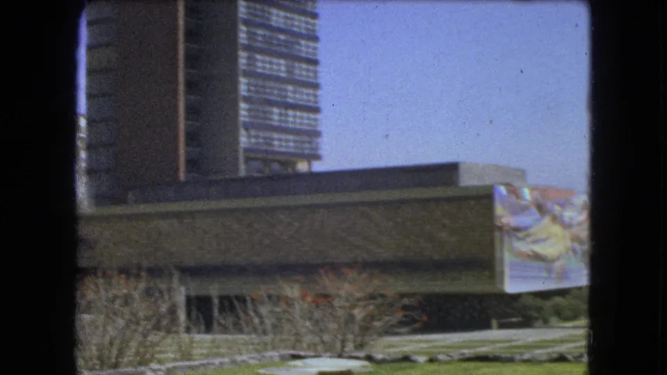 Mexico City venue exterior in 1962, showing a mural panel on a stone facade with lawns, paths, shrubs, and towers behind.