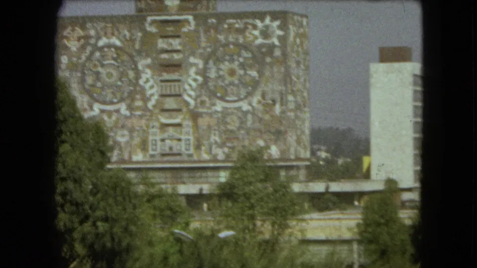 1962 Mexico City view of a large building facade with intricate mural patterns, partly obscured by trees and a dark foreground frame.