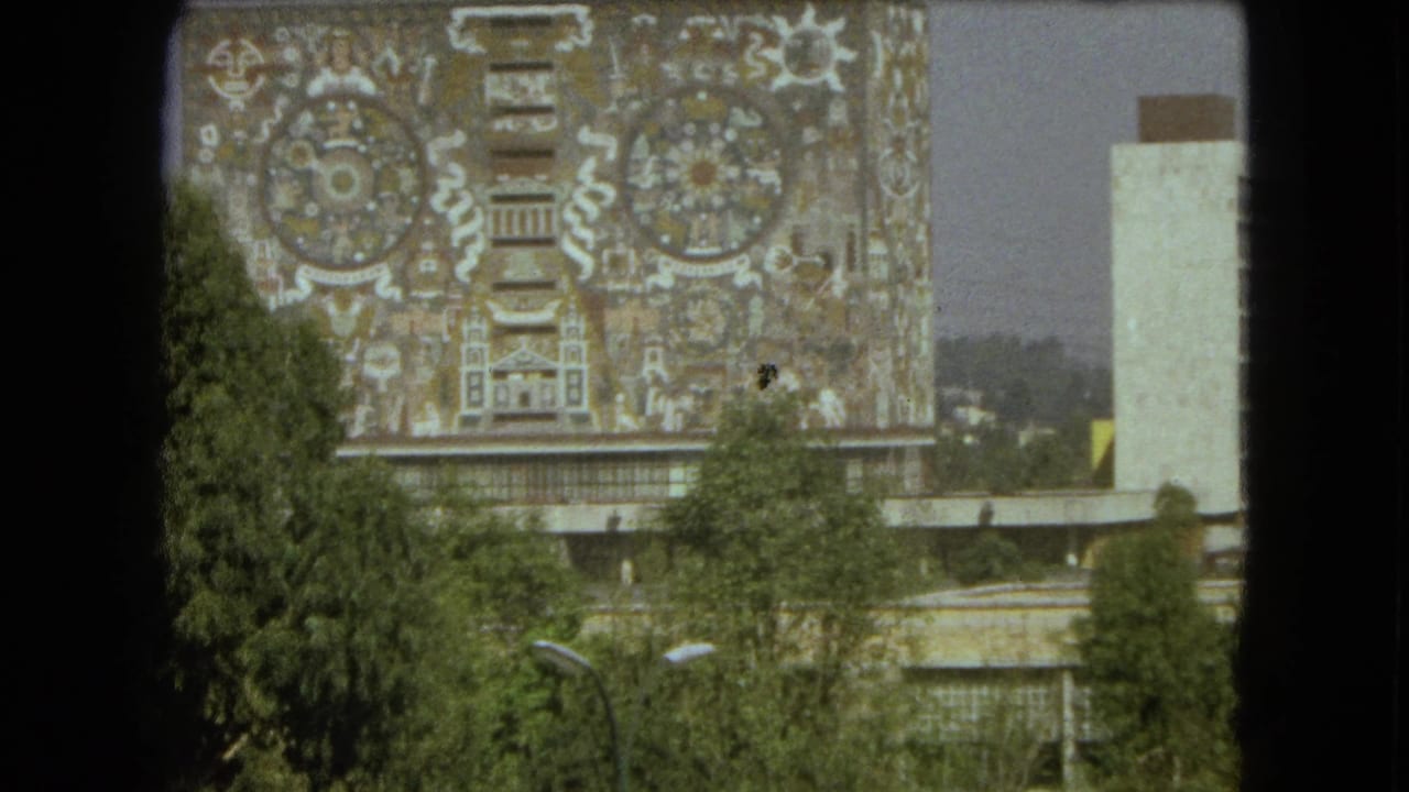 1962 Mexico City frame showing a monumental patterned mural facade, foreground trees, and a pale adjacent tower-like structure.