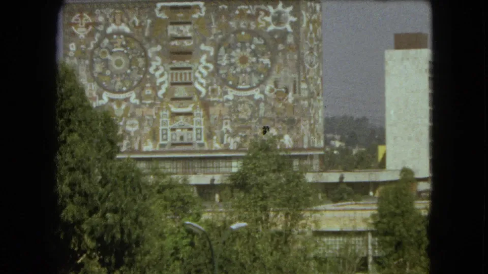 1962 Mexico City frame showing a monumental patterned mural facade, foreground trees, and a pale adjacent tower-like structure.