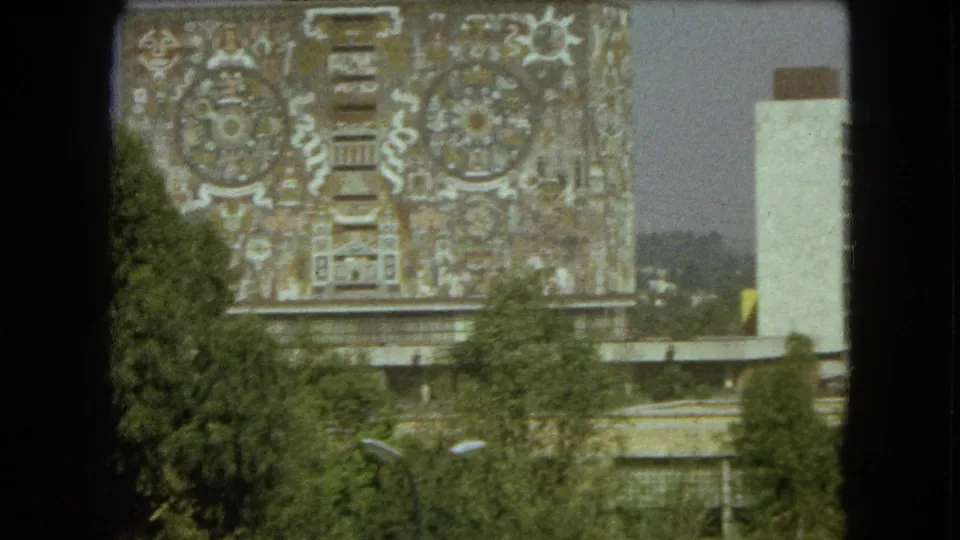 1962 Mexico City home-movie view of the mural-covered building, with trees low in the frame and dark film or window borders visible.
