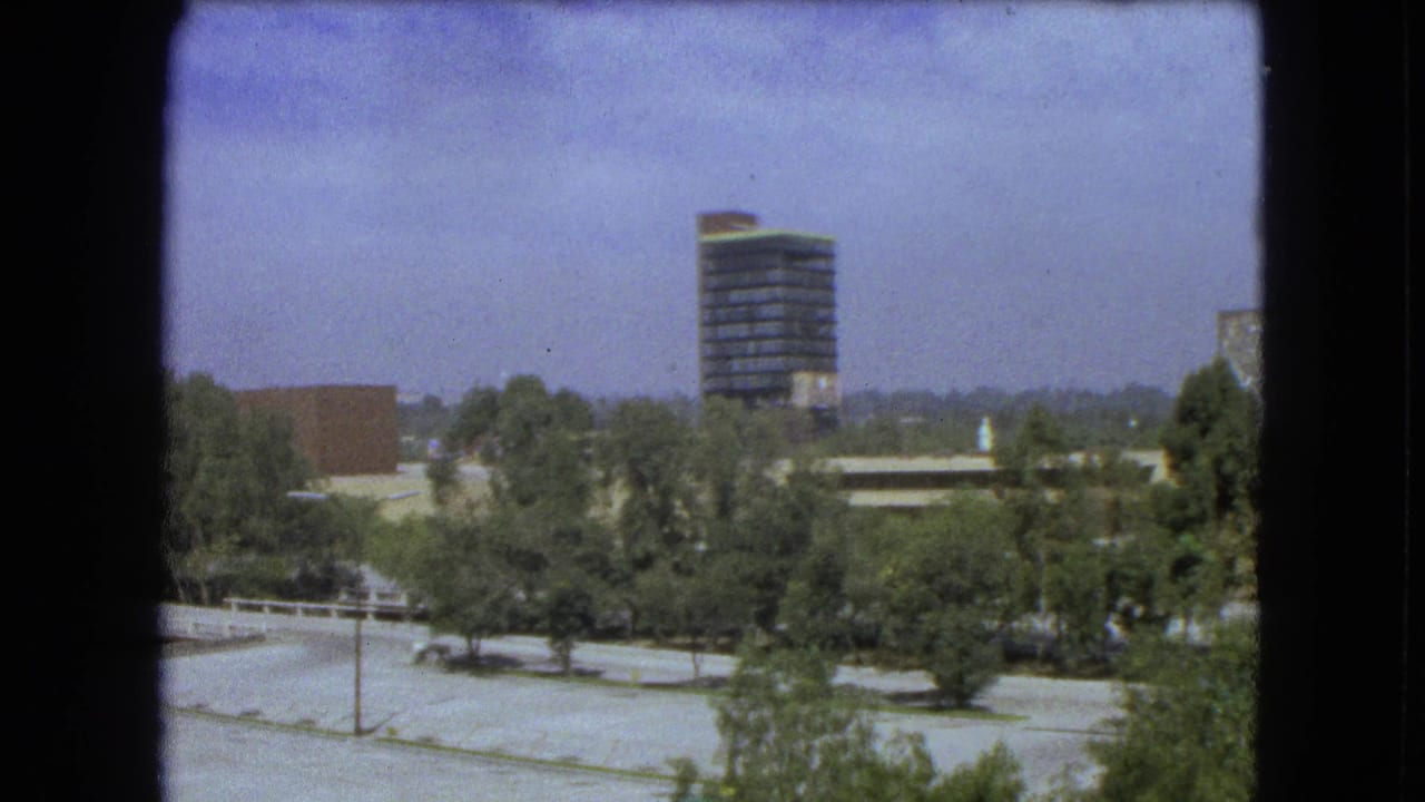 1962 Mexico City home-movie view from above with trees, paved areas, and a dark modern high-rise in the distance.