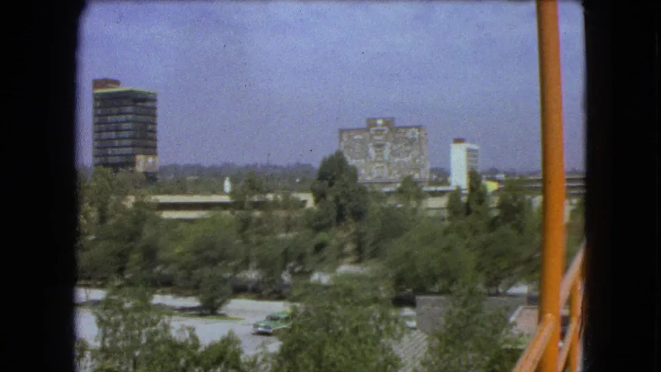 1962 Mexico City view with a high-rise, mural-covered building, street trees, parked cars, and an orange foreground pole.