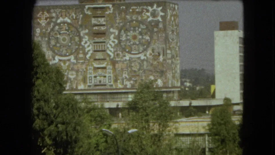 1962 Mexico City home-movie frame showing a large Aztec-inspired mural facade, pale tower, trees, and bright sky.