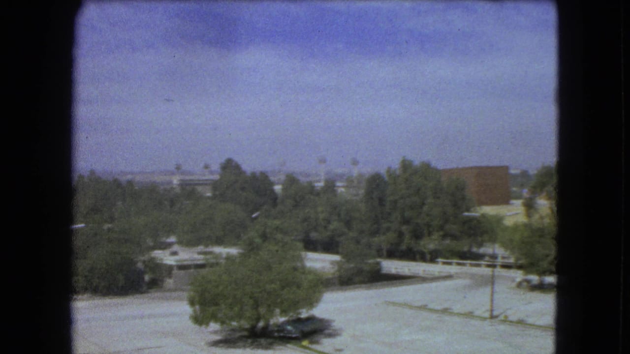 Panned view of trees, a paved lot, low buildings, and distant structures.