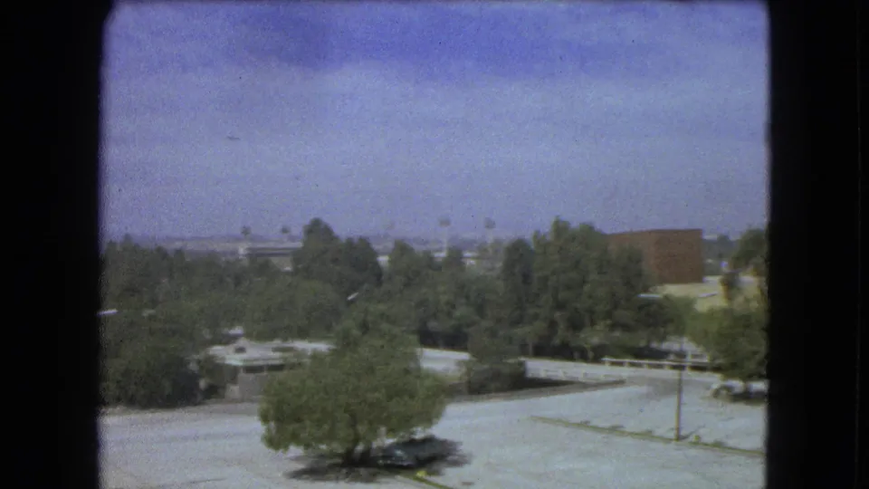 Elevated 1962 Mexico City view with leafy trees, pale paved lots, low commercial buildings, and distant urban forms.