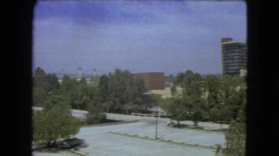 1962 Mexico City home-movie frame showing treetops, paved lots, a low brick building, and a modern high-rise tower at the right edge.