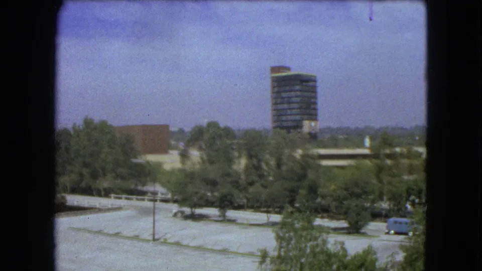 End of the 1962 Mexico City pan with trees, open paved space, low buildings, a modern tower, and a small blue vehicle near the lower right.