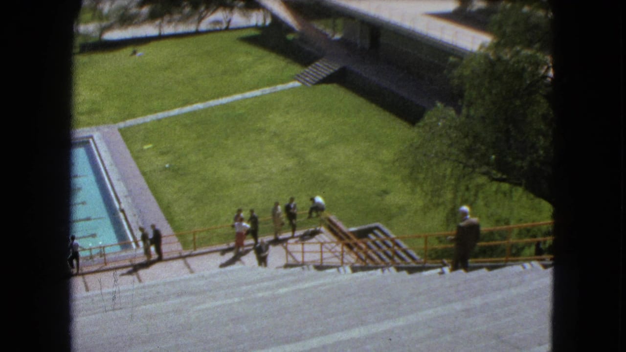 High view of a pool, stairs, lawn, and people beside a modernist campus building.
