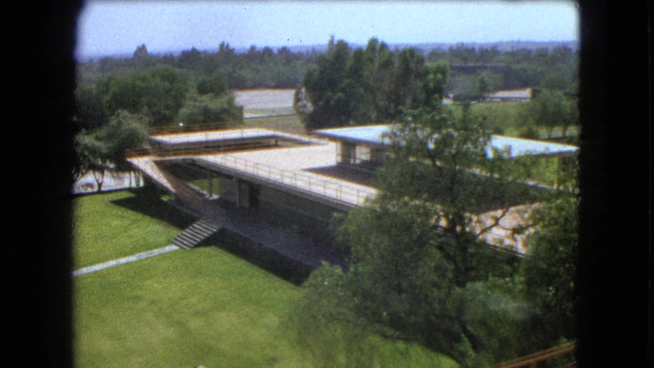 1962 Mexico City campus view through a dark frame, showing flat roofs, connecting walkways, lawns, and mature trees.