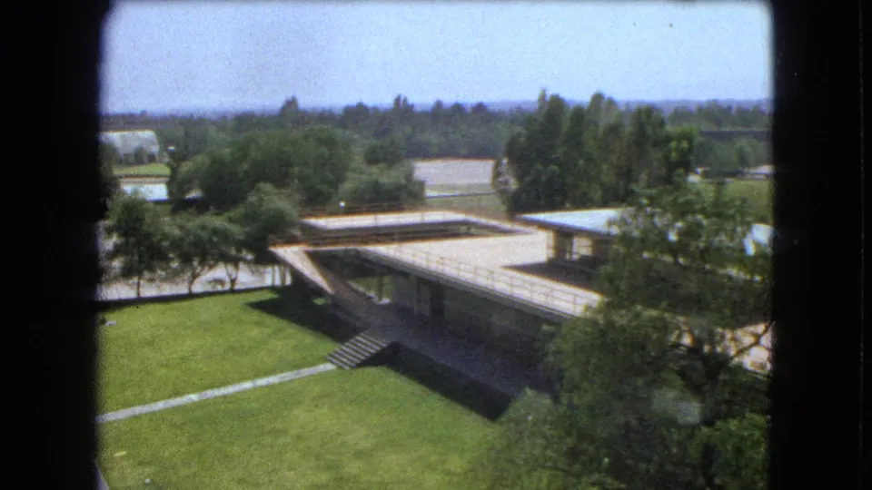 1962 Mexico City 8mm frame showing a low campus building, exterior stairs, open lawn, trees, and distant landscape.