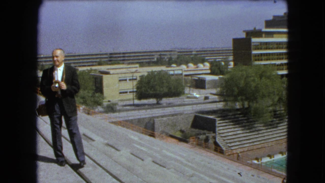 A suited man holds a camera on a concrete overlook in 1962 Mexico City, with roadways, trees, bleachers, and buildings behind him.