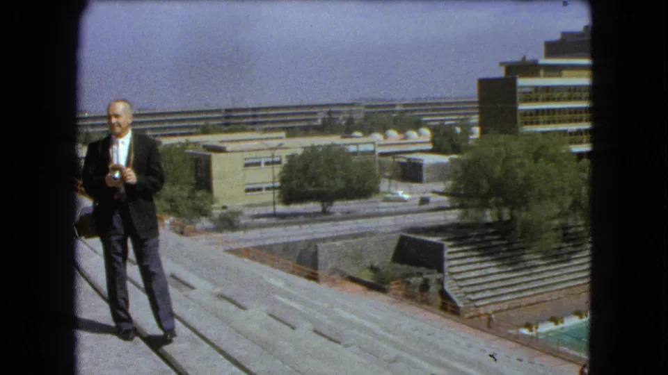 A suited man holds a camera on a concrete overlook in 1962 Mexico City, with roadways, trees, bleachers, and buildings behind him.