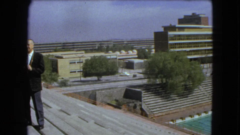 Partial 1962 Mexico City frame with a suited man at the edge, overlooking roadways, trees, bleachers, and modern buildings.