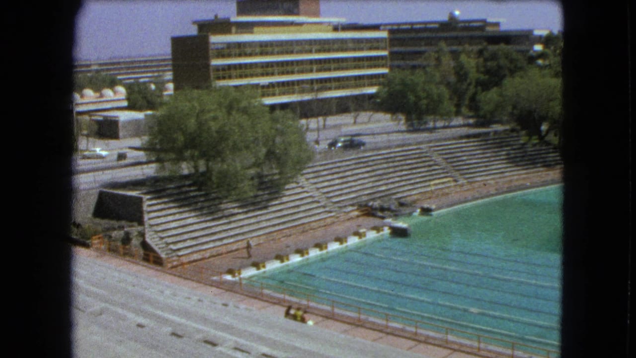 Outdoor swimming pool beside empty concrete bleachers, trees, roadway, and striped building.