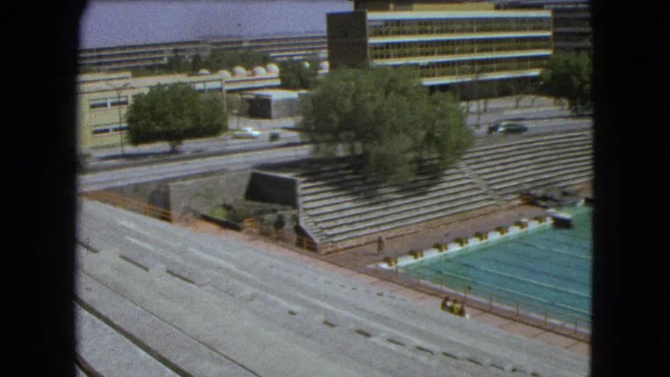 1962 Mexico City 8mm view across a broad road toward an outdoor pool, empty bleachers, trees, cars, and low institutional buildings.