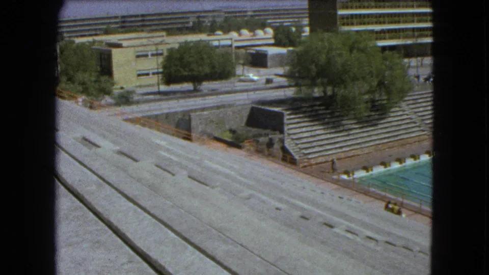 Final 1962 Mexico City frame showing a broad roadway beside the pool corner, stepped bleachers, trees, and striped building facade.