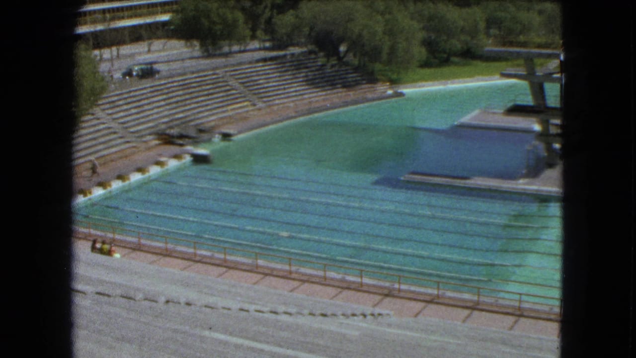 1962 Mexico City 8mm view of a mostly empty outdoor swimming stadium with turquoise lanes, concrete stands, trees, and a diving platform.