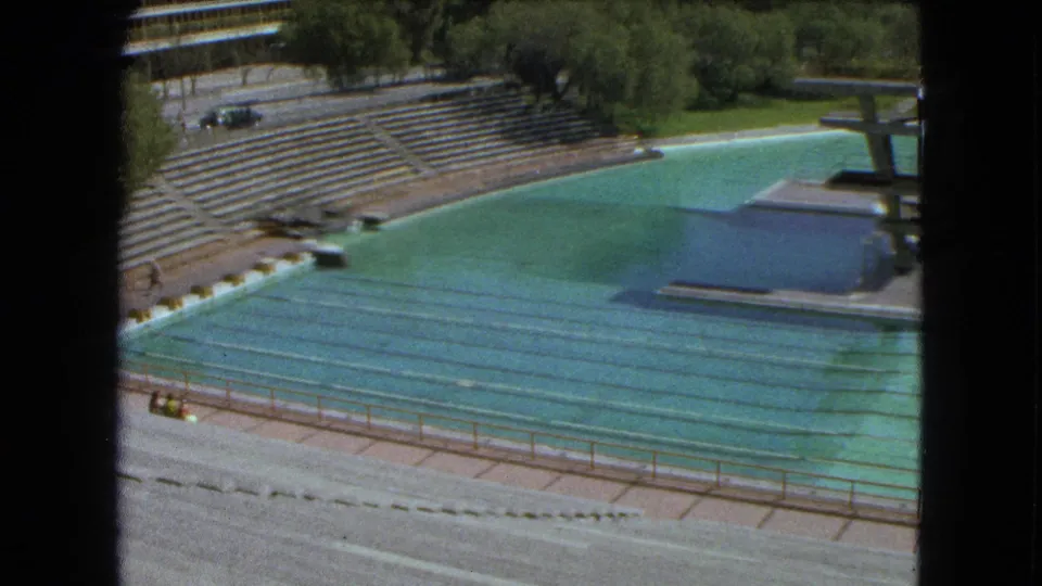 1962 Mexico City 8mm view of a mostly empty outdoor swimming stadium with turquoise lanes, concrete stands, trees, and a diving platform.