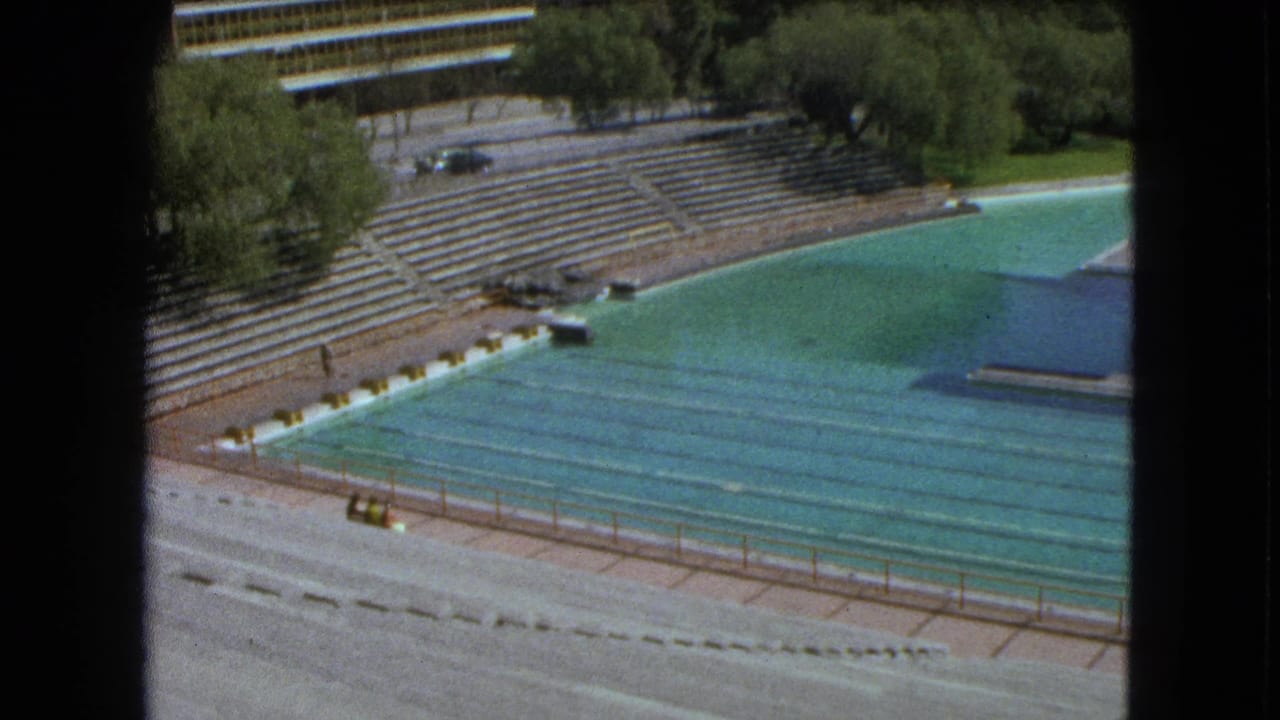 1962 Mexico City home-movie frame showing the outdoor pool, tiered bleachers, trees, and part of a diving platform in bright sun.