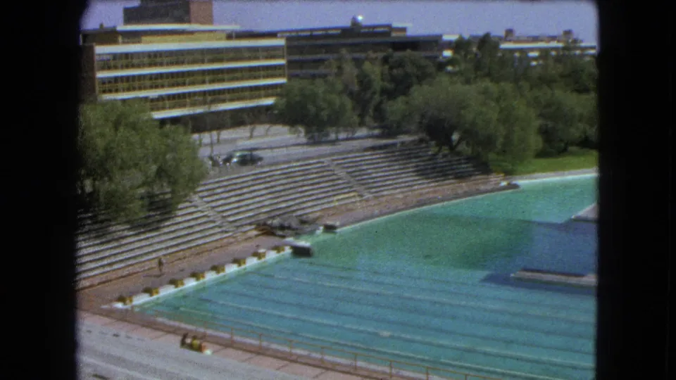 1962 Mexico City 8mm frame of a swimming stadium with turquoise water, stepped bleachers, trees, and a modern building in the background.