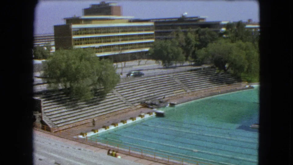 1962 Mexico City view across an outdoor swimming stadium toward tiered seating, trees, a few distant people, and a large building.