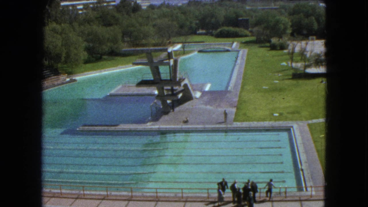 1962 Mexico City 8mm view of a large outdoor pool complex with lane markings, diving platforms, grass, trees, and distant people.