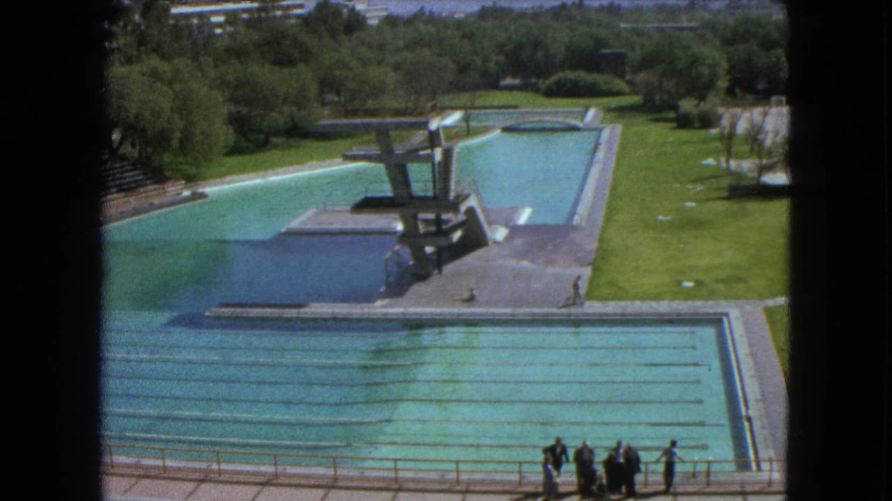 Outdoor pool complex with long lanes, diving structures, open grass, trees, and a small group near the foreground railing.
