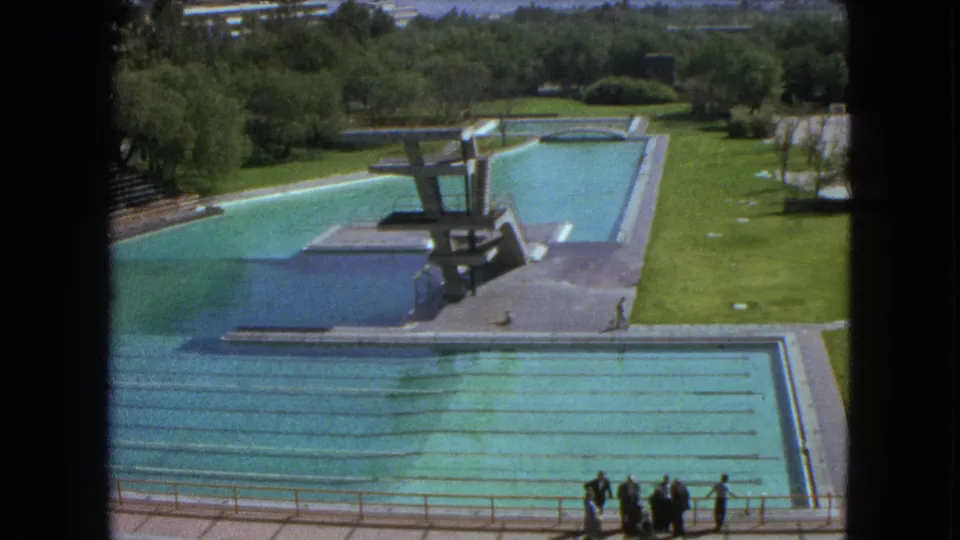 Elevated 1962 Mexico City pool view showing turquoise water, diving platforms, lawn, trees, and distant figures by the deck.