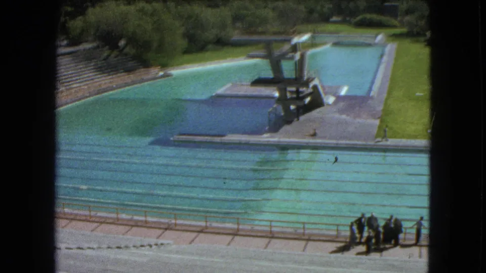 1962 Mexico City 8mm frame looking across an outdoor swimming pool toward diving platforms, trees, lawn, and a deckside group.