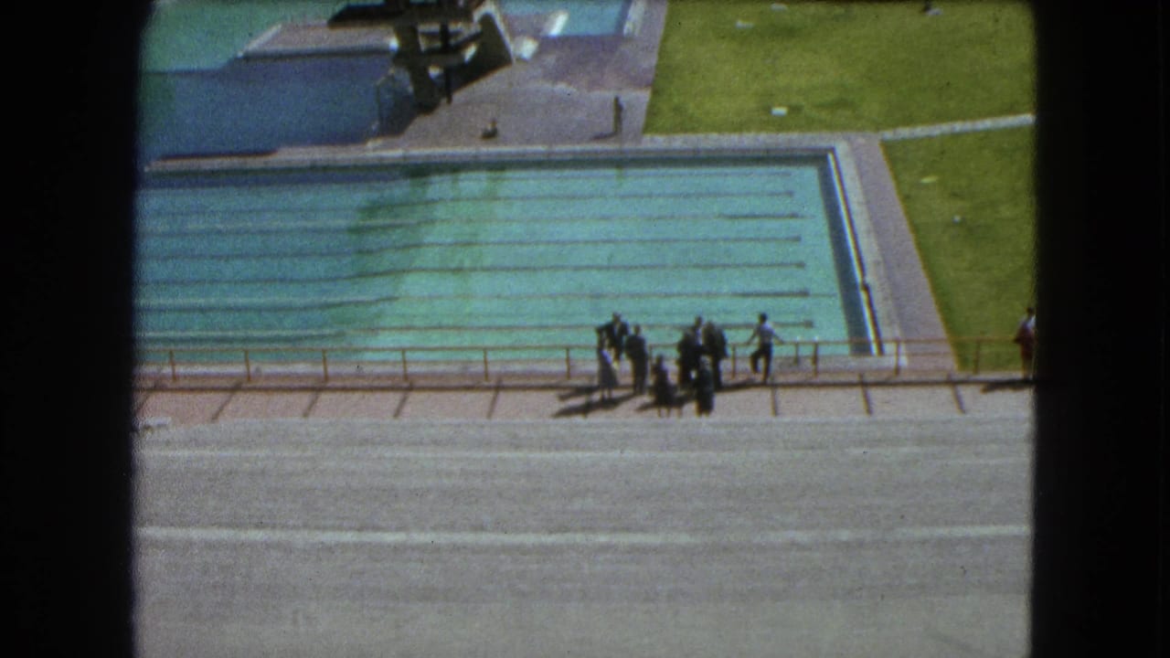 High-angle 1962 Mexico City view of a turquoise pool, poolside walkway, people near a railing, and grass around the complex.