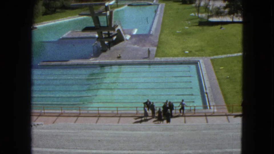1962 Mexico City 8mm frame showing the diving platform area, adjacent pools, lawn, walkways, and distant people at poolside.