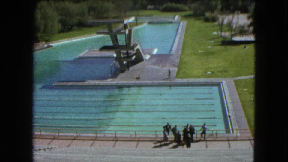 Wide 1962 Mexico City pool-complex view with a diving platform, lane-marked water, lawns, paths, and a small distant group.