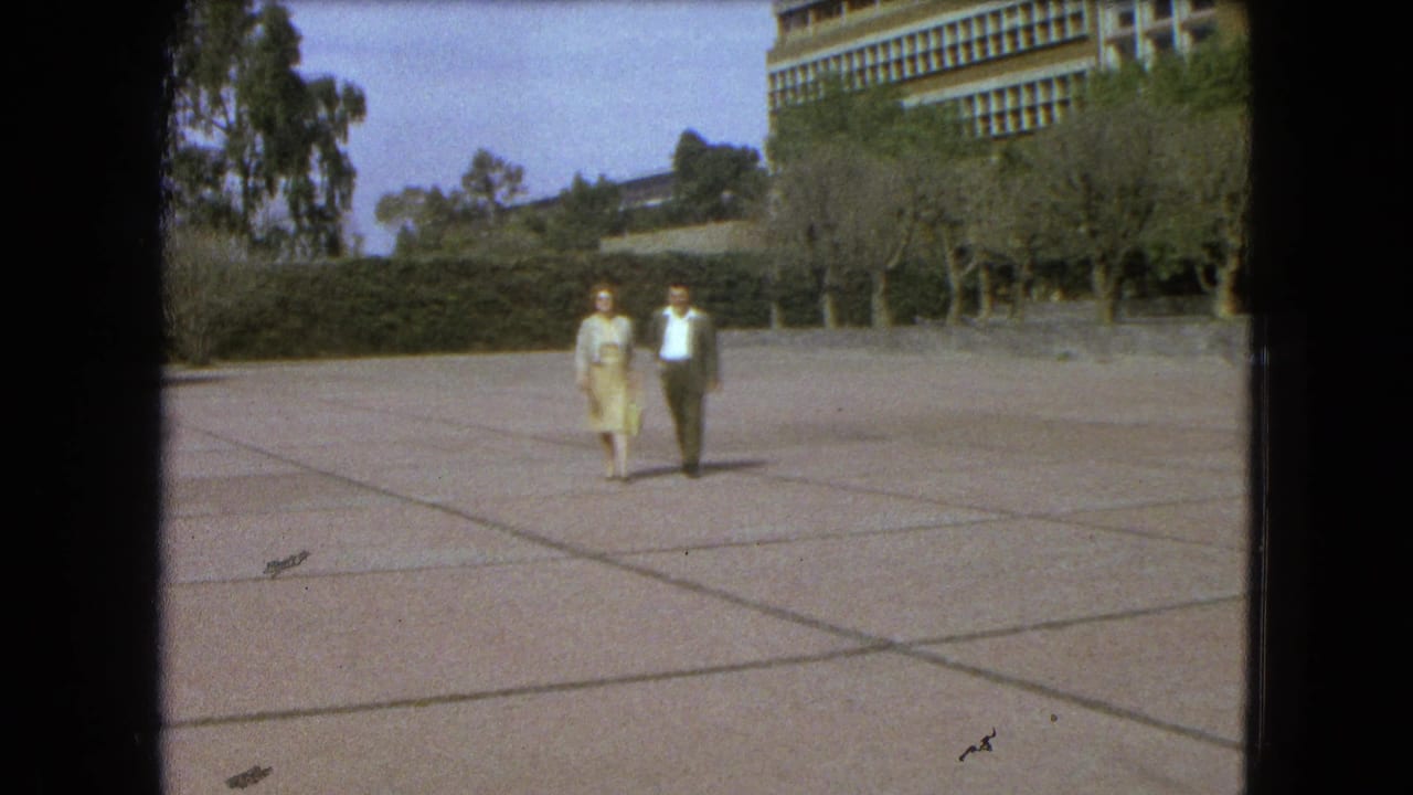 A woman and man walk across a broad paved plaza with trees and a large building behind them.