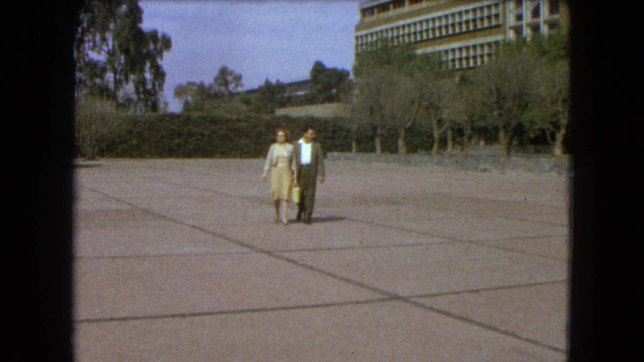 A woman in a light dress and a suited man walk side by side across a Mexico City esplanade in 1962 home-movie footage.