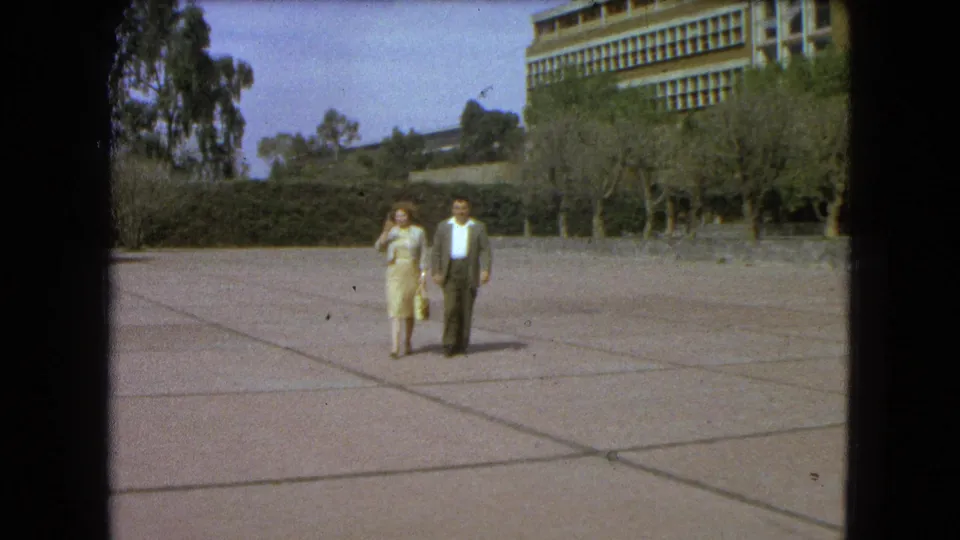 The couple continues toward the camera on a wide Mexico City plaza, with the woman raising a hand as the man walks beside her.