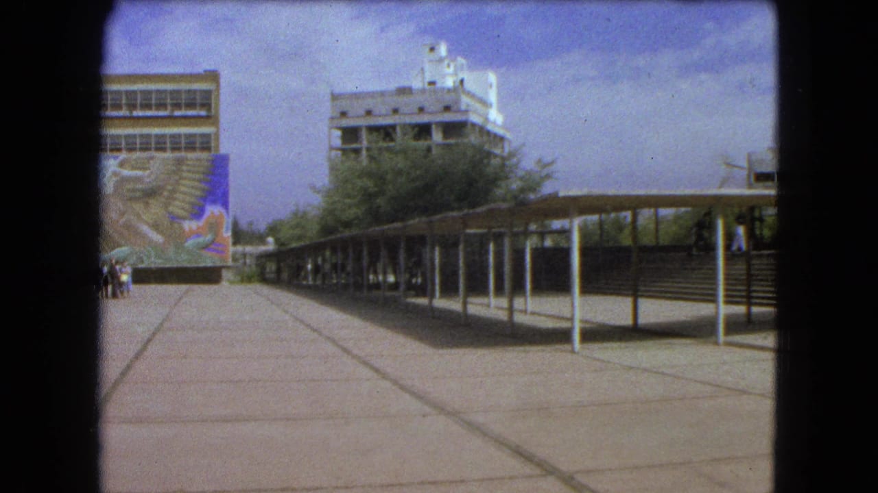 1962 Mexico City home-movie frame showing a sunlit plaza, modern buildings, a covered breezeway, and a colorful mural.