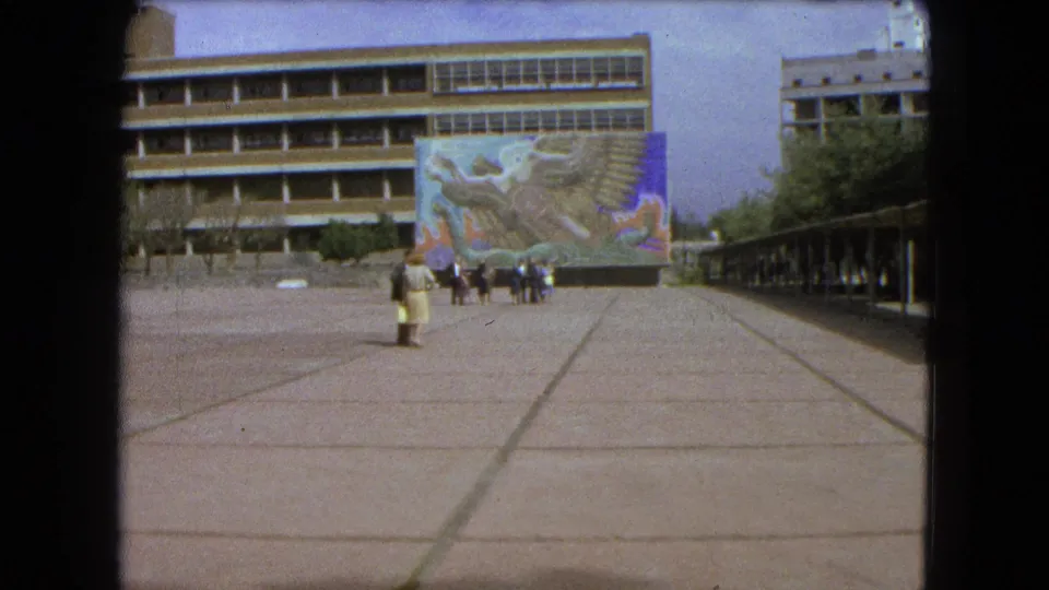 1962 Mexico City frame showing a tiled plaza, mural on a building facade, breezeway, and a foreground pedestrian in period clothing.