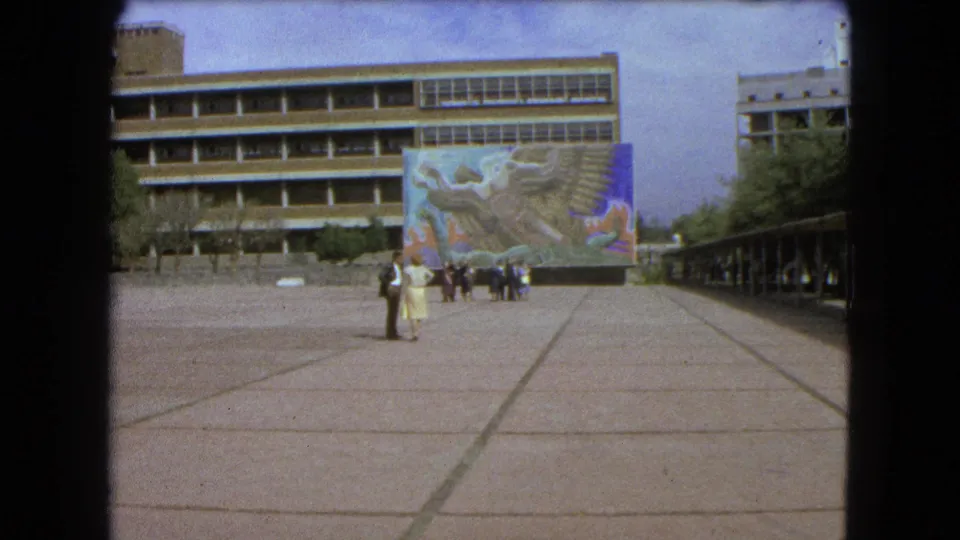 Mexico City home-movie frame from 1962 with pedestrians in a large plaza before a colorful mural and modern buildings.