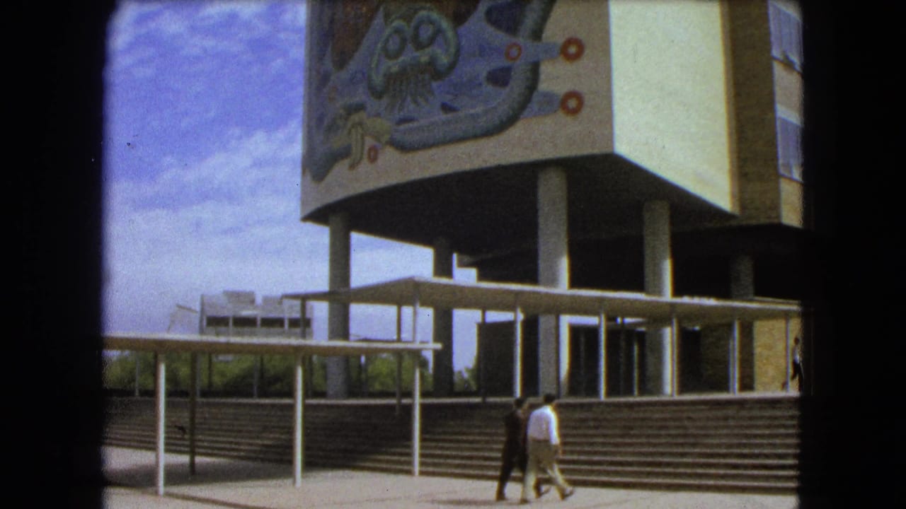 Two pedestrians walk past steps and covered railings below a muraled building facade.