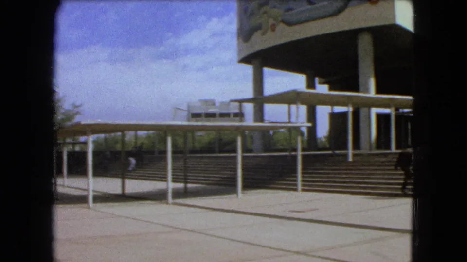 1962 Mexico City home-movie view across plaza paving toward covered walkways and a muraled modernist building.