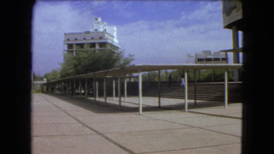 1962 Mexico City frame showing a broad public plaza with covered walkway, steps, trees, and modernist buildings.