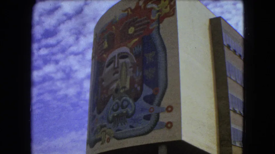 1962 Mexico City 8mm still showing the lower portion of a colorful facade mural on a modern building viewed from street level.